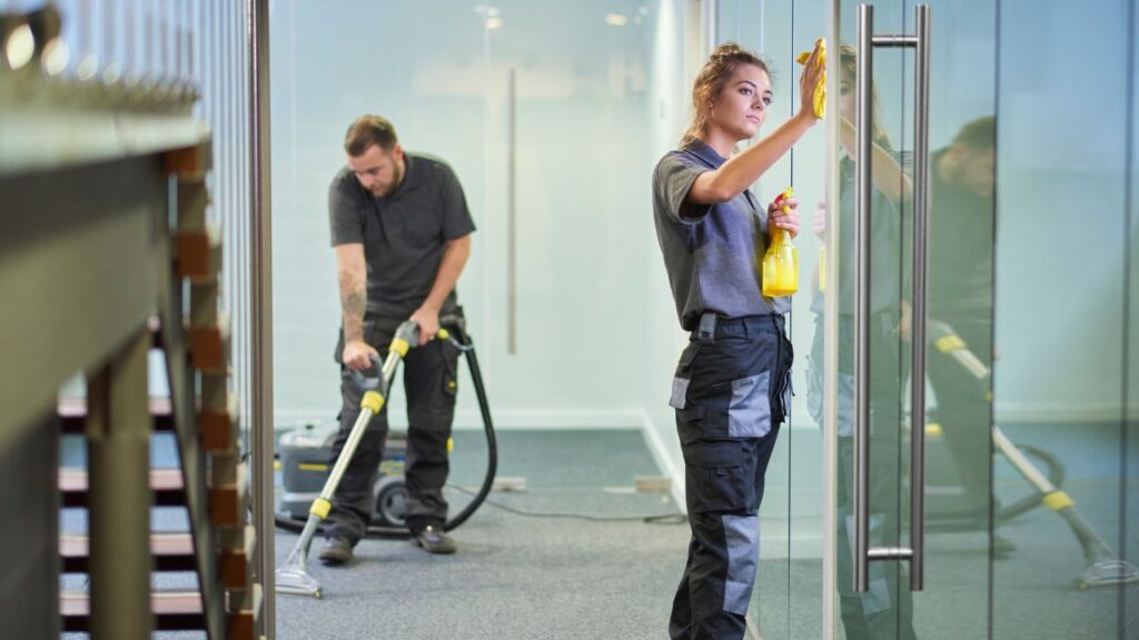 a man and a woman cleaning an office space with big windows and carpet floors