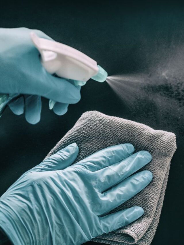 close up of hands wearing protection gloves, one hand holding a spray with a sanitizer and the other holding a cloth, sanitizin a black surface