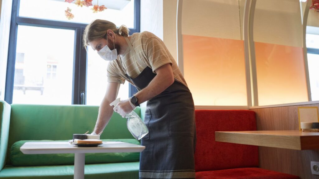 man wearing a cleaning apron, face maks and gloves, holding a sanitizer bottle while cleaning a table at a diner