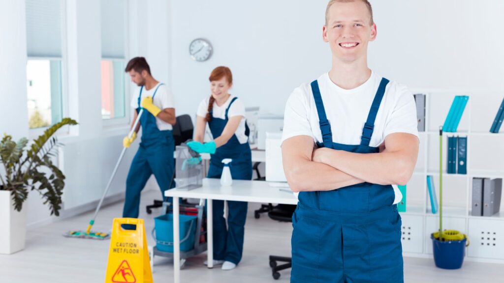 three professional cleaning staff organizing an office space with one, a white male staff, posing up front for a picture