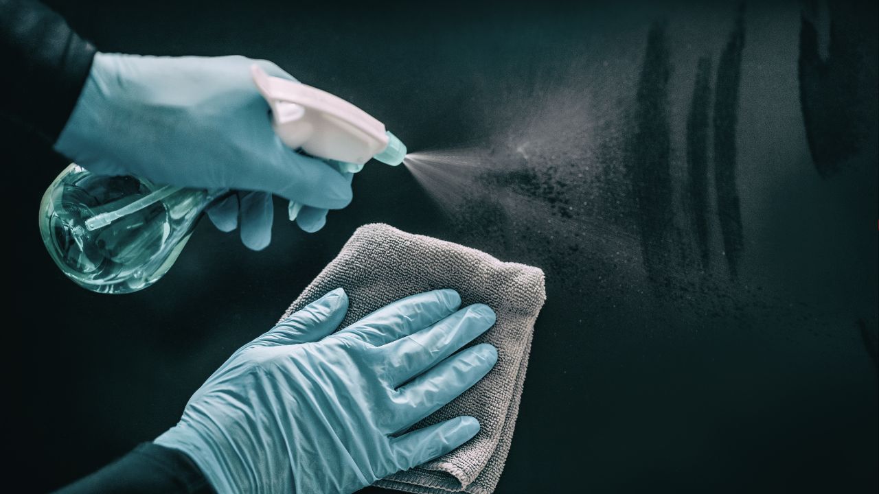 close up of hands wearing protection gloves, one hand holding a spray with a sanitizer and the other holding a cloth, sanitizin a black surface